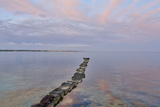 View of Saline dello Stagnone with a row of stones leading into the calm water under a pink sunset sky in Marsala Sicilia Italy.