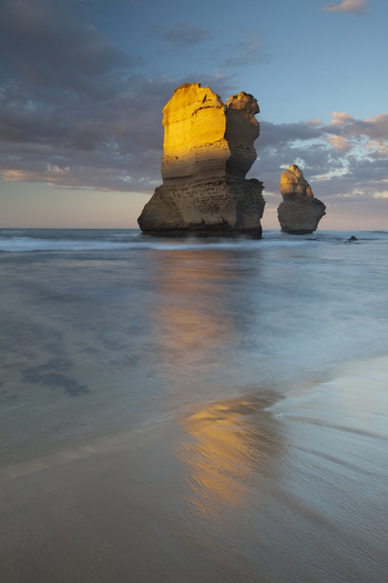 View of the Twelve Apostles limestone stacks illuminated by golden hour light with reflections on the wet sand and ocean waves in Australia. View of the Twelve Apostles limestone stacks illuminated by golden hour light with reflections on the wet sand and ocean waves in Australia.