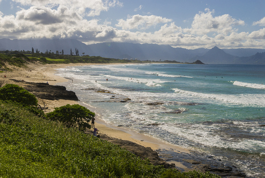 View of Kaneohe Bay with turquoise waves crashing on the sandy beach lush green vegetation in the foreground and mountains under a cloudy sky in Honolulu County Hawaii United States. View of Kaneohe Bay with turquoise waves crashing on the sandy beach lush green vegetation in the foreground and mountains under a cloudy sky in Honolulu County Hawaii United States.