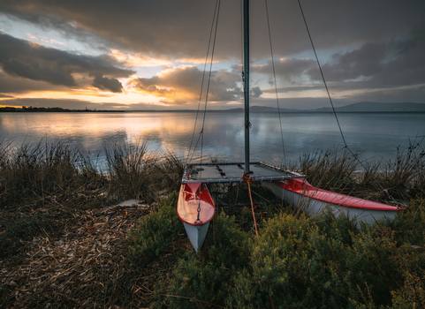 View of a red and white catamaran resting on the grassy shore of a calm bay at sunset with golden light reflecting on the water in Little Grove Western Australia Australia. View of a red and white catamaran resting on the grassy shore of a calm bay at sunset with golden light reflecting on the water in Little Grove Western Australia Australia.