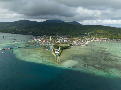 Aerial view of the vibrant town square meeting the tranquil turquoise sea contrasting with the lush green hills under a cloudy sky Karimunjawa Central Java Indonesia. Aerial view of the vibrant town square meeting the tranquil turquoise sea contrasting with the lush green hills under a cloudy sky Karimunjawa Central Java Indonesia.