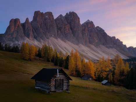 Aerial view of the Dolomites with rustic wooden huts nestled among vibrant autumn larch trees under a soft twilight sky in Italy.