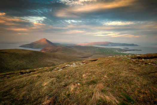 View of the rugged coastline and rolling hills of Keel West under a dramatic sunset sky with golden light hitting the peaks in County Mayo Ireland. View of the rugged coastline and rolling hills of Keel West under a dramatic sunset sky with golden light hitting the peaks in County Mayo Ireland.