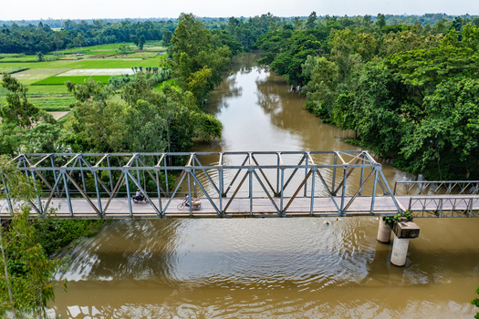 Aerial view of a metal truss bridge over a muddy river with a cyclist and motorcyclist crossing surrounded by lush trees and fields in Bogura Rajshahi Division Bangladesh. Aerial view of a metal truss bridge over a muddy river with a cyclist and motorcyclist crossing surrounded by lush trees and fields in Bogura Rajshahi Division Bangladesh.