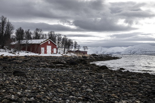 View of a traditional red wooden cabin on a rocky shoreline with snow covered mountains and a cloudy sky Tromso Troms Norway.