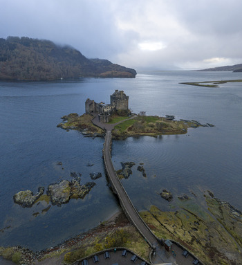 Aerial view of Eilean Donan Castle on a small island connected by a stone bridge over Loch Duich under a cloudy sky in Dornie Scotland United Kingdom.