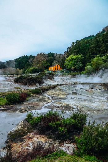 View of geothermal landscape with steaming vents and an orange house nestled amongst lush greenery a stark contrast to the grey earth Furnas Azores Portugal.