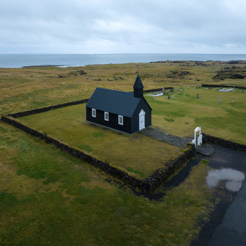 Aerial view of the iconic black church embraced by a stone wall amidst the expansive green landscape meeting the moody sea B&uacute;&eth;ir Sn&aelig;fellsb&aelig;r Iceland.