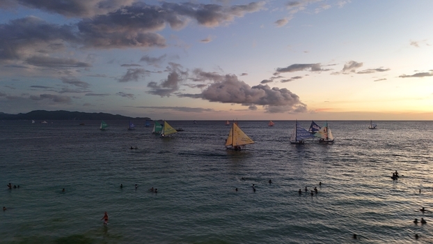 Malay Philippines 02 May 2026 Aerial view of Boracay Island with traditional paraw sailboats on the calm sea at sunset featuring people swimming and a cloudy sky.