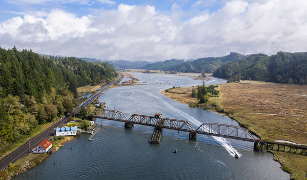 Aerial view of the Siuslaw River Train Bridge spanning the river with a boat leaving a wake surrounded by green hills wetlands and a highway in Florence Oregon United States.