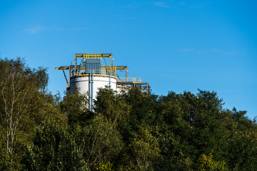 View of an industrial storage tank with a yellow metal framework on top surrounded by dense green trees under a clear blue sky in Poland.