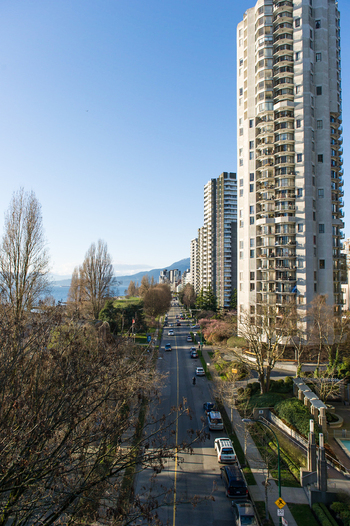 View of high rise residential buildings along a street with trees and parked cars under a clear blue sky in Vancouver Canada.