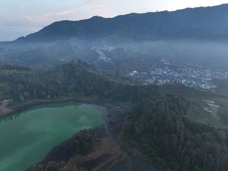 Aerial view of Telaga Warna lake in the Dieng Plateau surrounded by lush green hills misty mountains and the village of Kejajar Central Java Indonesia. Aerial view of Telaga Warna lake in the Dieng Plateau surrounded by lush green hills misty mountains and the village of Kejajar Central Java Indonesia.