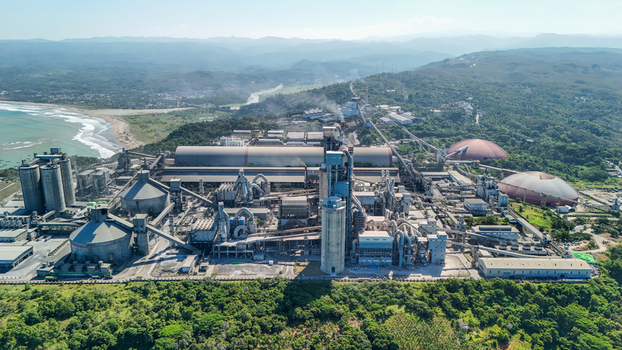 Aerial view of a large industrial cement plant complex near the coastline featuring tall silos storage domes and manufacturing facilities surrounded by green hills in Indonesia.