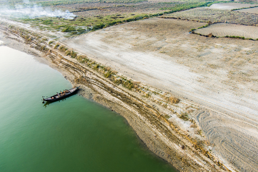 Aerial view of a small wooden boat with people moored on the sandy riverbank of the Padma River with smoke rising in the distance in Rajshahi Rajshahi Division Bangladesh.