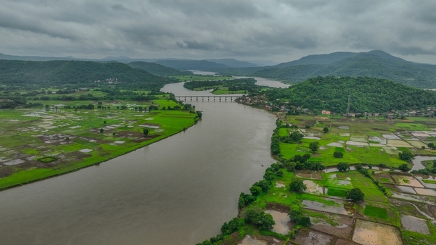 Aerial view of the Mumbai Goa Highway bridge crossing a winding river through lush green agricultural fields and hills in Mahad Maharashtra India.