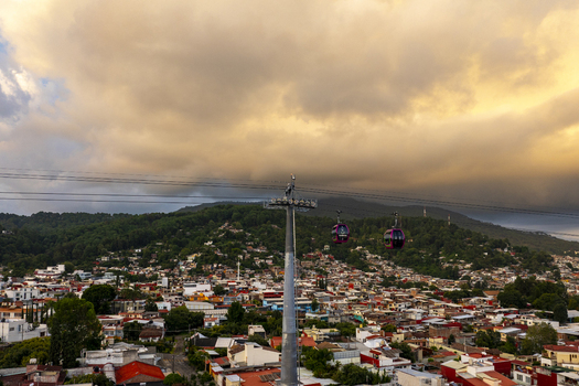Aerial view of the cable car teleferico Uruapan gliding over the dense urban landscape with colorful rooftops lush green hills and a dramatic golden hour sky in Uruapan Michoacan Mexico.