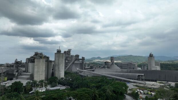 Aerial view of a large industrial cement factory with tall silos and conveyor belts surrounded by lush greenery under a cloudy sky in Klapanunggal West Java Indonesia. Aerial view of a large industrial cement factory with tall silos and conveyor belts surrounded by lush greenery under a cloudy sky in Klapanunggal West Java Indonesia.