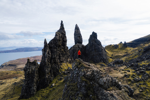 Aerial view of The Storr with a person in a red jacket standing on a rocky outcrop surrounded by dramatic basalt pinnacles and rugged terrain in Portree Scotland United Kingdom. Aerial view of The Storr with a person in a red jacket standing on a rocky outcrop surrounded by dramatic basalt pinnacles and rugged terrain in Portree Scotland United Kingdom.