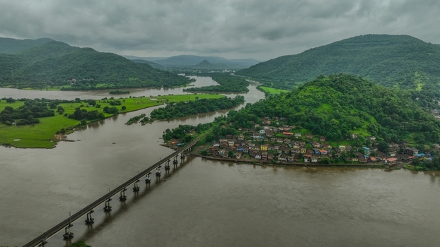 Aerial view of the Mumbai Goa Highway bridge crossing a wide river with a small village nestled at the base of lush green hills in Mahad Maharashtra India.