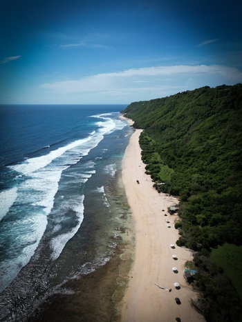 Aerial view of Pantai Nunggalan featuring a long sandy beach turquoise ocean waves and lush green cliffs under a clear blue sky in Kecamatan Kuta Selatan Bali Indonesia.