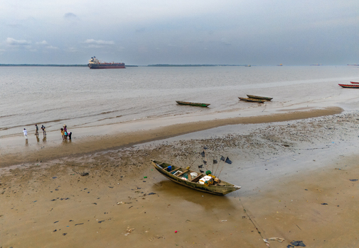 Aerial view of Bonny Island showing traditional wooden fishing boats on the sandy shore with people standing nearby and a large cargo ship in the distance at Bonny Town Rivers Nigeria. Aerial view of Bonny Island showing traditional wooden fishing boats on the sandy shore with people standing nearby and a large cargo ship in the distance at Bonny Town Rivers Nigeria.