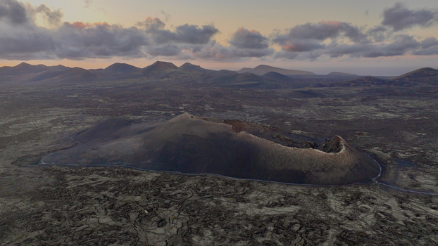 Aerial view of a volcanic crater surrounded by vast lava fields and distant mountains under a soft sunset sky in Lanzarote. Aerial view of a volcanic crater surrounded by vast lava fields and distant mountains under a soft sunset sky in Lanzarote.