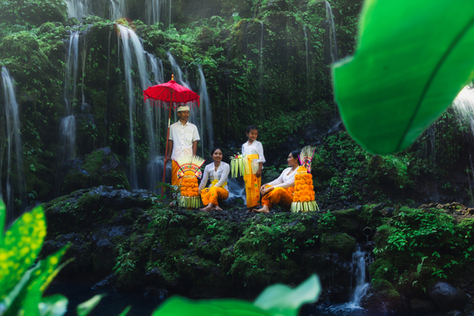 Bali Indonesia View of people adorned in vibrant orange and white nestled against a cascading waterfall s lush green backdrop under the soft shade of a red umbrella. Bali Indonesia View of people adorned in vibrant orange and white nestled against a cascading waterfall s lush green backdrop under the soft shade of a red umbrella.