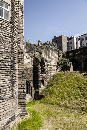 View of the historic stone walls and fortifications of Newcastle Castle with grassy embankments and modern buildings in the background in Newcastle upon Tyne England United Kingdom. View of the historic stone walls and fortifications of Newcastle Castle with grassy embankments and modern buildings in the background in Newcastle upon Tyne England United Kingdom.