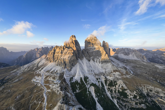 Aerial view of the majestic Tre Cime di Lavaredo peaks pierce the sky their rugged faces kissed by golden sunlight contrasting with the deep shadows Tre Cime di Lavaredo Belluno Italy. Aerial view of the majestic Tre Cime di Lavaredo peaks pierce the sky their rugged faces kissed by golden sunlight contrasting with the deep shadows Tre Cime di Lavaredo Belluno Italy.