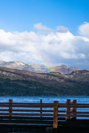 View of Parque Nacional Los Alerces with a wooden pier in the foreground a calm lake and mountains under a blue sky with a faint rainbow in Futaleufu Department Chubut Province Argentina. View of Parque Nacional Los Alerces with a wooden pier in the foreground a calm lake and mountains under a blue sky with a faint rainbow in Futaleufu Department Chubut Province Argentina.