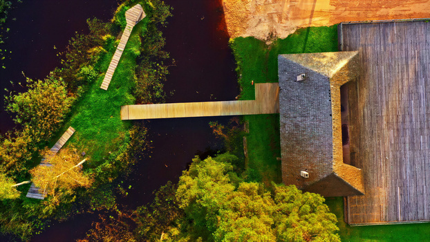 Aerial view of a wooden building with a large deck and a wooden walkway extending over a dark pond surrounded by lush green trees and vegetation in Latvia. Aerial view of a wooden building with a large deck and a wooden walkway extending over a dark pond surrounded by lush green trees and vegetation in Latvia.