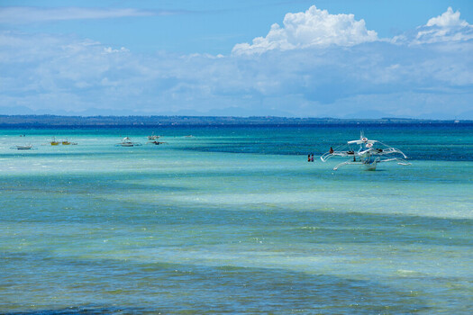 View of traditional outrigger boats floating in the turquoise waters of Bantayan Island with people swimming nearby under a bright blue sky in Cebu Central Visayas Philippines. View of traditional outrigger boats floating in the turquoise waters of Bantayan Island with people swimming nearby under a bright blue sky in Cebu Central Visayas Philippines.