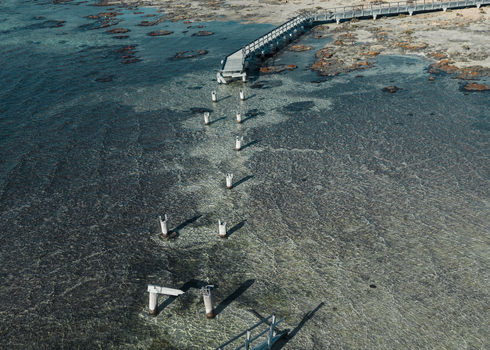 Aerial view of a wooden jetty extending into the clear turquoise waters of Shark Bay with visible sand patterns and submerged pilings in Denham Western Australia Australia. Aerial view of a wooden jetty extending into the clear turquoise waters of Shark Bay with visible sand patterns and submerged pilings in Denham Western Australia Australia.