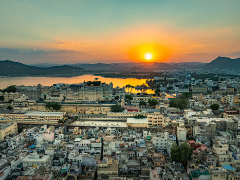 Aerial view of the City Palace complex overlooking Lake Pichola at sunset with historic rooftops and surrounding hills in Udaipur Rajasthan India.