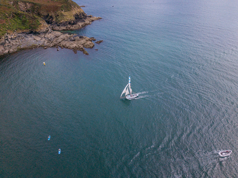 Aerial view of a sailboat navigating the Fowey river near the rocky coastline with small boats and calm blue water in Fowey Cornwall United Kingdom. Aerial view of a sailboat navigating the Fowey river near the rocky coastline with small boats and calm blue water in Fowey Cornwall United Kingdom.