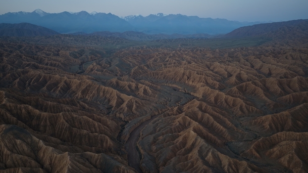 Aerial view of the rugged arid canyons and deep gullies of the Issyk Kul landscape with snow capped mountains in the distance in Issyk Kul Region Kyrgyzstan. Aerial view of the rugged arid canyons and deep gullies of the Issyk Kul landscape with snow capped mountains in the distance in Issyk Kul Region Kyrgyzstan.
