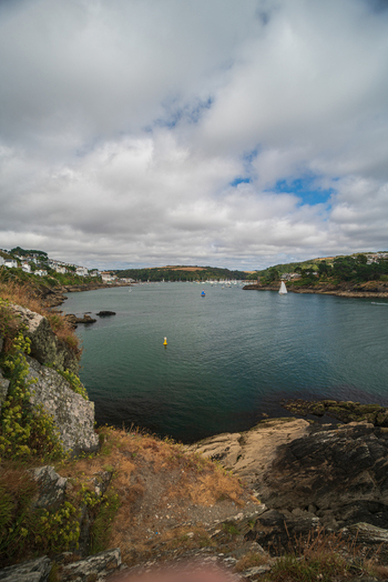 View of the Fowey River with boats moored near the town of Fowey rocky cliffs in the foreground and a cloudy sky in Fowey Cornwall United Kingdom. View of the Fowey River with boats moored near the town of Fowey rocky cliffs in the foreground and a cloudy sky in Fowey Cornwall United Kingdom.