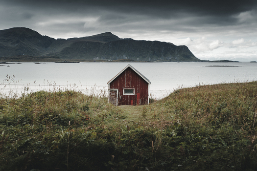 View of a traditional red wooden cabin situated on a grassy field overlooking a calm bay with mountains in the background under a cloudy sky in Ramberg Nordland Norway. View of a traditional red wooden cabin situated on a grassy field overlooking a calm bay with mountains in the background under a cloudy sky in Ramberg Nordland Norway.