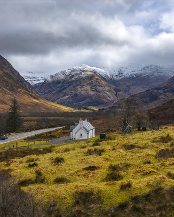 Aerial view of a white cottage nestled in a vast valley surrounded by snow capped mountains and dramatic clouds Scotland United Kingdom. Aerial view of a white cottage nestled in a vast valley surrounded by snow capped mountains and dramatic clouds Scotland United Kingdom.