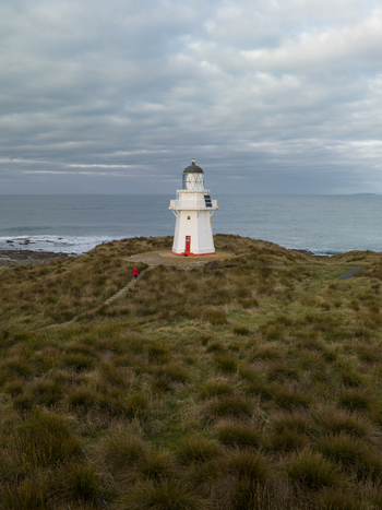 Aerial view of the stark white lighthouse a beacon against the moody grey sea and sky standing sentinel on a grassy knoll Otara Southland Region New Zealand.