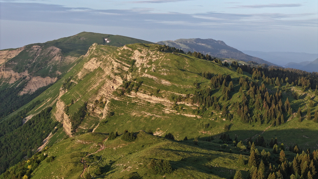 Aerial view of Col de la Faucille mountain pass with lush green slopes rocky ridges and coniferous forests under soft morning light Gex Auvergne Rhone Alpes France.