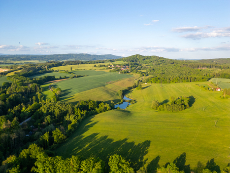 Aerial view of the rolling green hills and fields of the Cesky Raj rural landscape with a small pond and patches of forest under a clear sky in Czechia.