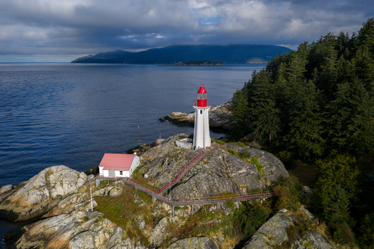 Aerial view of Point Atkinson Lighthouse on a rocky headland with a red roofed building surrounded by evergreen forest and the blue waters of the Pacific Ocean West Vancouver British Columbia Canada.