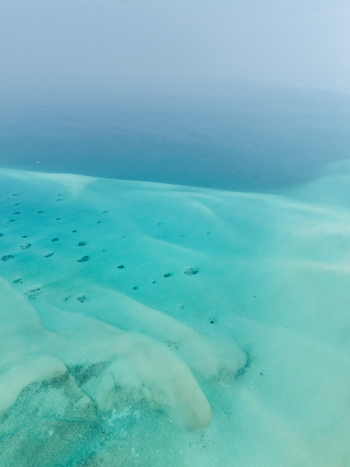 Aerial view of vibrant turquoise shallows revealing sandy patterns and dark coral heads transitioning into the deep hazy blues of the ocean horizon Thuraakunu Thiladhunmathee Uthuruburi Maldives. Aerial view of vibrant turquoise shallows revealing sandy patterns and dark coral heads transitioning into the deep hazy blues of the ocean horizon Thuraakunu Thiladhunmathee Uthuruburi Maldives.