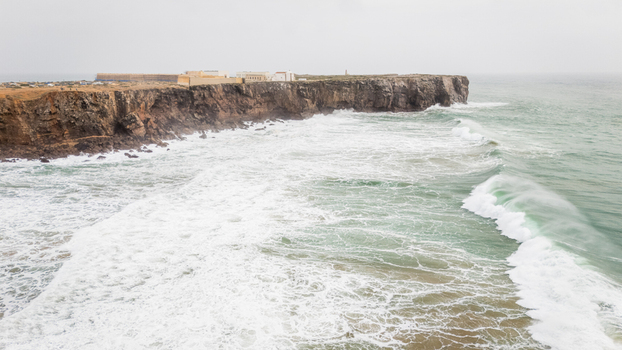 Aerial view of the historic Sagres Fortress perched on rugged cliffs with powerful waves crashing against the shore under an overcast sky in Sagres Faro Portugal. Aerial view of the historic Sagres Fortress perched on rugged cliffs with powerful waves crashing against the shore under an overcast sky in Sagres Faro Portugal.