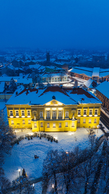 Aerial view of the illuminated Museum of Srem building surrounded by snow covered grounds and rooftops during a winter evening in Sremska Mitrovica Vojvodina Serbia.