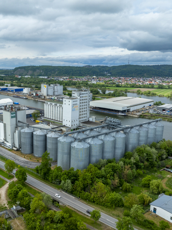 Aerial view of large industrial grain silos and warehouses with the BayWa logo situated along a river canal with lush green trees and hills in the background.
