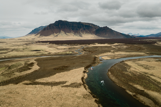 Aerial view of a person kitesurfing in a winding river through the vast tundra landscape with a large mountain in the background in Borgarbyggd Iceland. Aerial view of a person kitesurfing in a winding river through the vast tundra landscape with a large mountain in the background in Borgarbyggd Iceland.