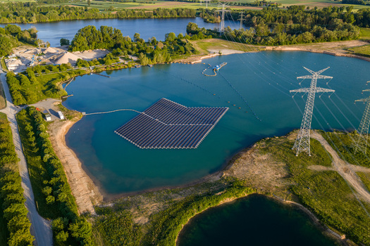Aerial view of floating solar farm on a blue lake surrounded by lush green trees and power pylons in Bensheim Hessen Germany. Aerial view of floating solar farm on a blue lake surrounded by lush green trees and power pylons in Bensheim Hessen Germany.